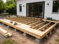 Grundstomme för altan byggs. Träbjälkar på betongplintar, redo att bygga altan. Verktyg ligger redo.