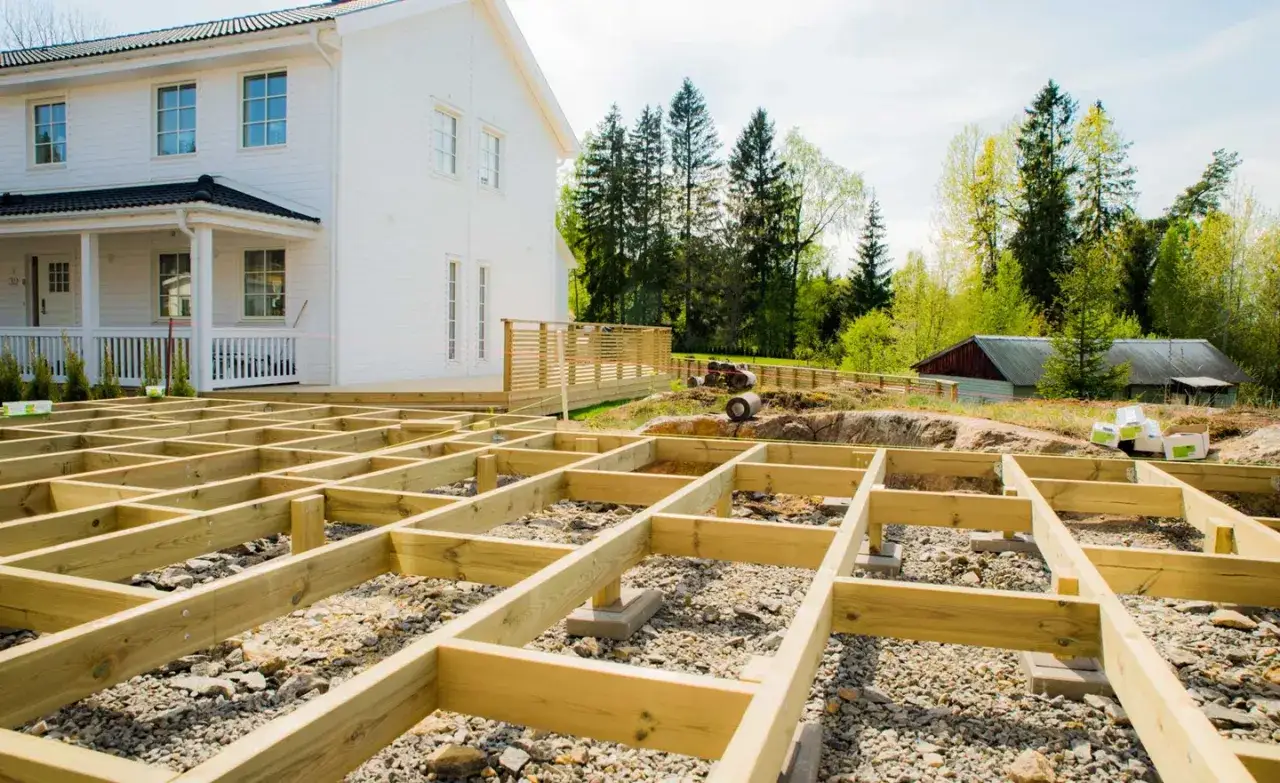 Grundstomme för att bygga altan vid ett vitt hus. Träbjälkar ligger redo för att skapa en ny uteplats.